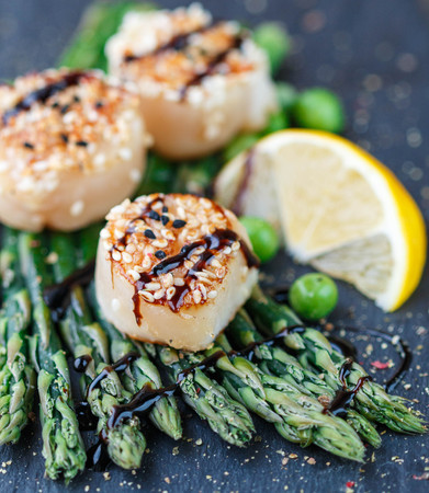 Fried scallop  with sesame seeds and balsamic sauce, asparagus, lemon and green peas on a black plateの写真素材