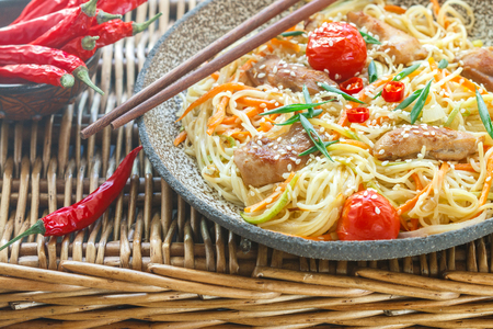 Stir fried noodle with chicken, vegetable (carrot, tomato,  zucchini, garlic, red pepper, green onion), sesame seeds and soy sauce on clay plate close up. Asian cuisine.  Selective focusの写真素材