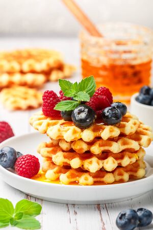 Viennese crispy waffles with fresh berries (raspberries and blueberries) with honey and mint leaves on a white plate on the table. Delicious homemade summer gourmet Breakfast. Rustic style. Selective focusの写真素材
