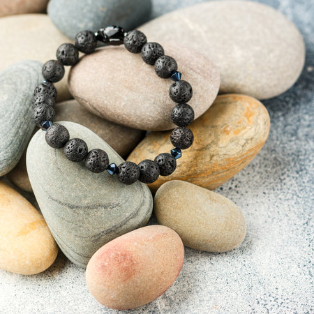 Beautiful bracelet made of natural black volcanic lava. Jewelry made of round-shaped beads is placed on large sea stones. Selective focus, square pictureの写真素材