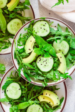 Green vegan or vegetarian salad of fresh vegetables-arugula, cucumber, avocado, green peas, coriander, basil with lemon juice and black sesame. Three servings on a gray background. Healthy food, dietの写真素材
