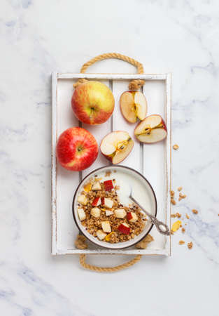 Homemade granola (muesli) with oatmeal, nuts, flaxseed, cornflakes, fresh red apple, cinnamon and yogurt. Delicious healthy breakfast on a white plate on a marble background. Diet. Proper nutrition. Top view, copy spaceの写真素材