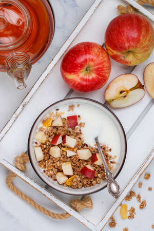 Homemade granola (muesli) with oatmeal, nuts, flaxseed, cornflakes, fresh red apple, cinnamon and yogurt. Delicious healthy breakfast on a white plate on a marble background. Diet. Proper nutrition. Top viewの写真素材