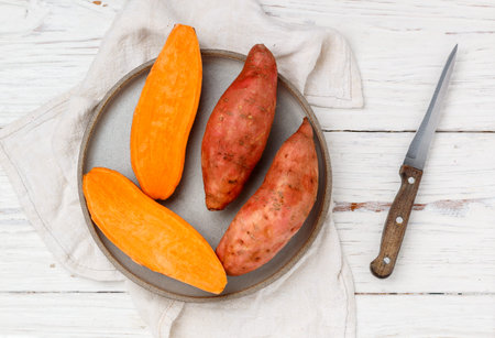 Raw organic whole and sliced sweet potatoes in a gray plate on a white wooden background. Fresh Root vegetables. Selective focus, top view and copy spaceの写真素材