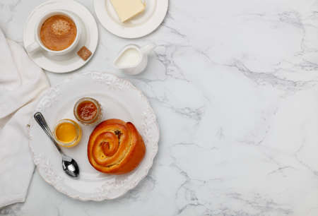 A sweet bun with raisins, jam and honey in a white plate on a marble background for breakfast. Coffee, milk, butter on the table. Selective focus, top view and copy spaceの写真素材