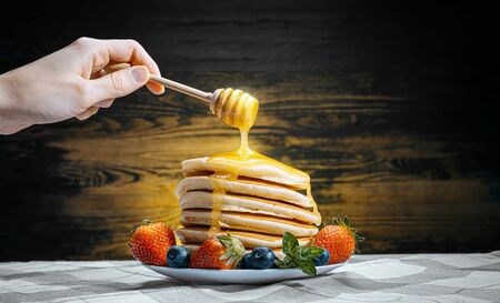 on classic homemade pancakes with berries on a white saucer on a dark wooden background in a rustic style pours delicious honey.の写真素材