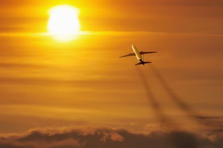 Airliner heads off in the direction of the setting sun against orange cloudy sky.の写真素材