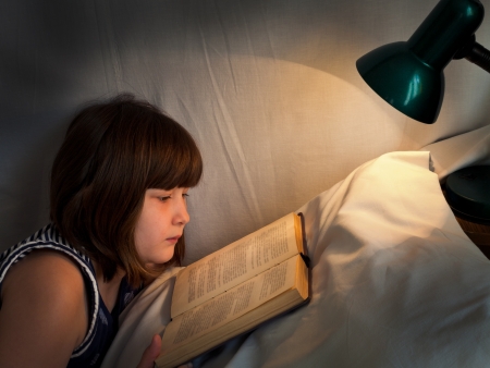 teen girl reading book on bed at night by light of lamp on bedの写真素材