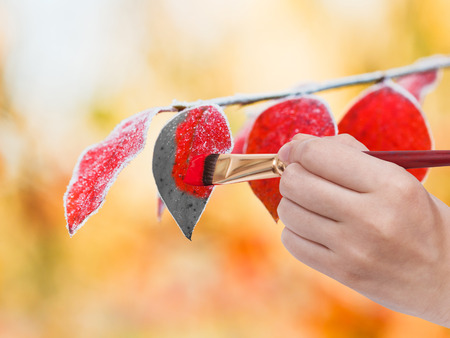nature concept - hand with paintbrush paints frozen leaves in red colour in cold autumn dayの写真素材