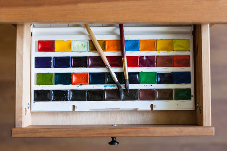 above view of watercolor paints set and painting brushes in open drawer of nightstand