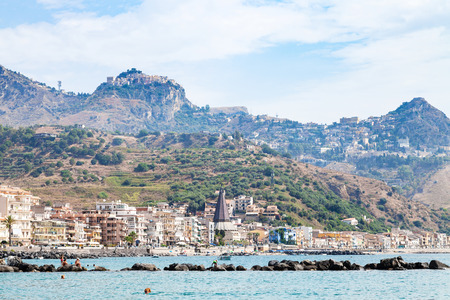 GIARDINI NAXOS, ITALY - JUNE 28, 2017: people on beach near waterfront of Giardini-Naxos town and view of Taormina city on cape. Giardini Naxos is seaside resort on Ionian Sea coast since the 1970sのeditorial素材