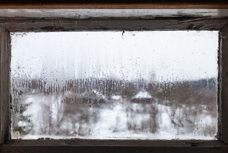 blurry view of russian village through water drops from melting ice on surface of frozen window of rural house in cold winter day (focus on surface of the glass)の写真素材