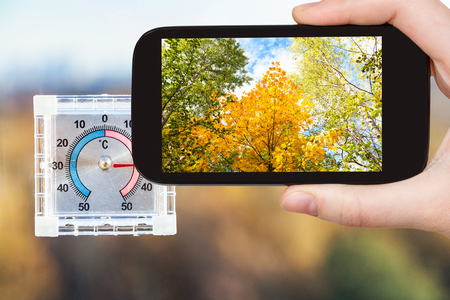 travel concept - tourist photographs yellow trees and outdoor thermometer on home window in hot autumn day on smartphone in Moscow, Russiaの写真素材