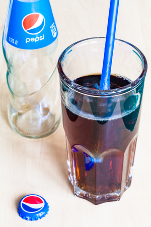 MOSCOW, RUSSIA - APRIL 4, 2019: Pepsi soft drink in glass with blue drinking tube, used crown cap and empty glass bottle on wooden table. Pepsi is carbonated soft drink manufactured by PepsiCoのeditorial素材