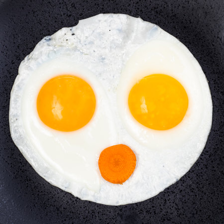 fried eggs and slice of carrot on black plate close up. Fried eggs like face with open mouthの写真素材