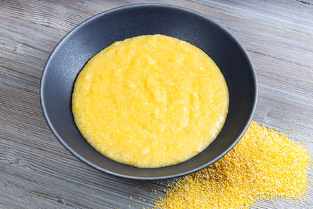 cooked maize porridge in gray bowl and handful of cornmeal on gray wooden tableの写真素材