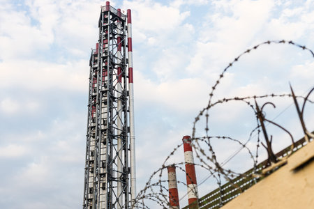 factory chimneys against blue sky with white clouds behind yellow fence with barbed wire (focus on...