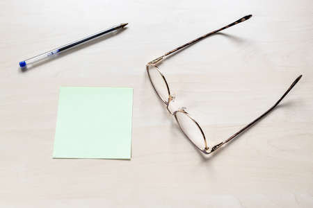 top view of eyeglasses, plastic blue pen on blank green sheet of note paper on light brown wooden boardの写真素材