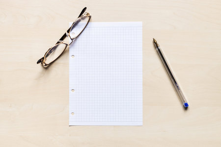top view of eyeglasses, pen and blank sheet of squared paper on light brown wooden board