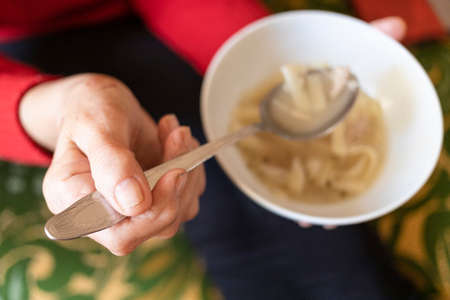 top view of hand of old woman holding tablespoon of chicken noodle soup (focus on the upper foreground thumb)の写真素材