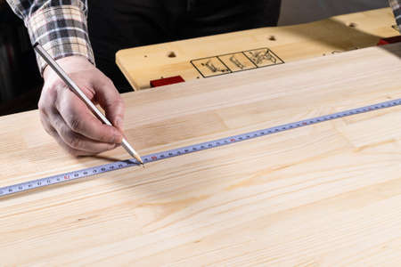 hand of carpenter with pencil marks size on wooden board with measuring tape on workbench at homeの写真素材