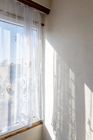 tulle curtains on home window and wall lit by sun and view of urban houses in background