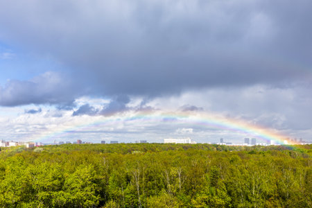 gray rainy clouds and rainbow in blue sky over green city park after rain on sunny spring dayの写真素材