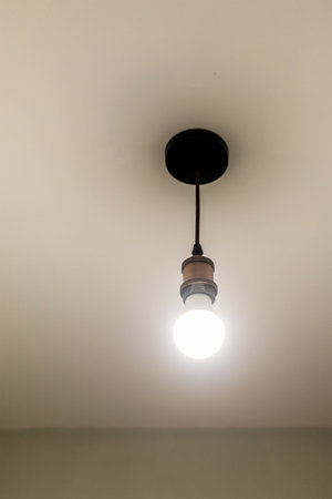 lit LED light bulb in brass electrical socket hangs on electrical wires from ceiling in room during...