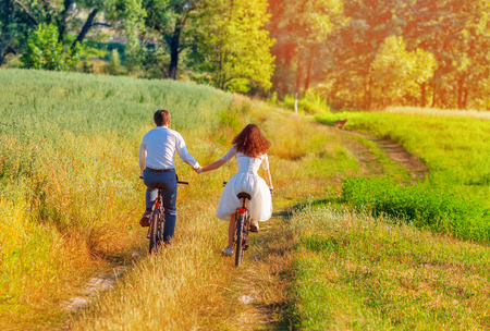 Save to a lightbox  Find Similar Images  Share Stock Photo: Young happy bride and groom ride bicycles in the meadow back to camera and holding handsの写真素材
