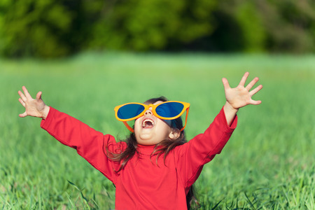 Happy little girl wearing big sunglasses with hands up enjoying sun in the fieldの写真素材