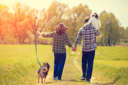 Happy family with dog walking on the rural dirt road on the field. Little girl sitting on dad's shoulder. Woman and man holding hands. Woman keeps her dog on a leash. Family and dog back to cameraの写真素材