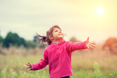 Happy little girl with hands up on the meadow in sunny dayの写真素材