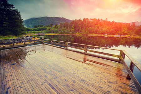 Wooden deck on a lakeshore. Rocky shore of the mountain lake in the rainy autumn morning. Beautiful nature Norway. Reflection on the water.の写真素材