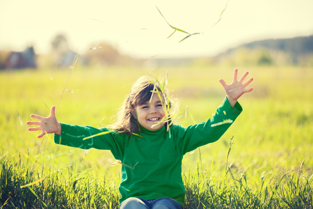 Happy laughing little girl with hands in the air sitting on the field in sunny dayの写真素材