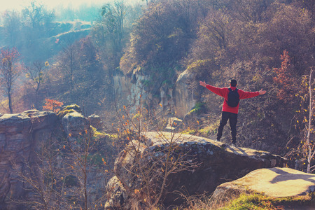A man with hands in the air standing on a rock over a beautiful canyon on a sunny autumn day. Man looking at the canyonの写真素材