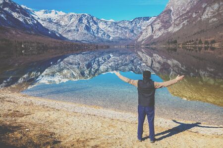 Man with hands in the air standing on the shore of the mountain lake in early spring. Bohinj lake, Slovenia, Europeの写真素材