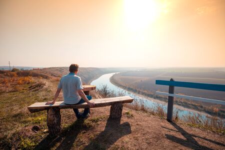 Mountain landscape with a river in the valley. A man sits on a bench on a mountain and looks at a beautiful landscape at sunsetの写真素材