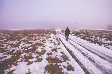 A man walks in the countryside on a foggy winter day. Dirt road among the fields covered with the first snowの写真素材