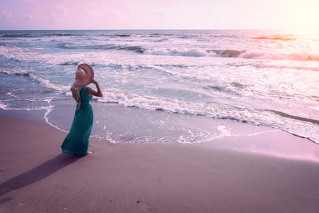 Seascape during pink sunrise. Woman on the beach. A young happy woman in a long green dress and straw hat walks on the seashore. The girl looks at the magical sunriseの写真素材
