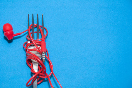 Red Earphones on metal fork around classic blue background. Broken headphones wrapped around a fork....