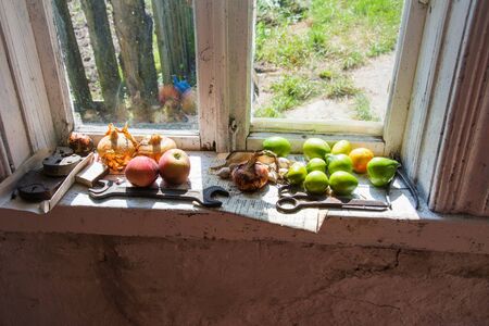 Rural still life. On the old white wooden windowsill are apples, onions, green tomatoes, matches, scissors, pliers, a wrench, padlocks, an old newspaperの写真素材