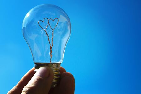 Young man holds a light bulb with two hearts inside on blue sky background for copyspace. The concept of love, loyalty, strong family, unity. Close up.の写真素材