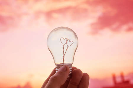 Man hold light bulb in hand, inside which there are two hearts against the sky and the sun.の写真素材
