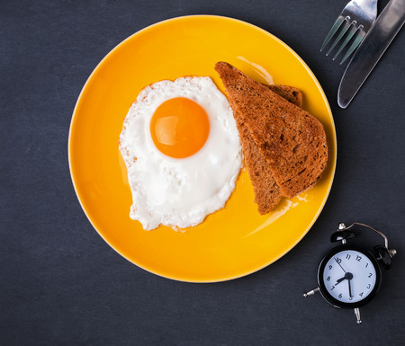 Fried egg and bread on a yellow plate and alarm clock on the black background, top viewの写真素材