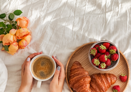 Womans hands holding a cup of coffee on the bed with white bedsheet.の写真素材