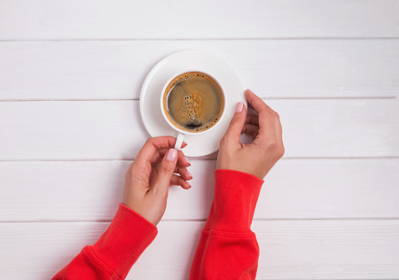 Womans hands in red clothing holding a cup of coffee on the white wooden tableの写真素材