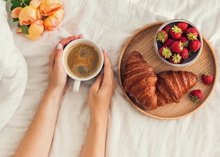 Womans hands holding a cup of coffee on the bed with white bedsheet.の写真素材