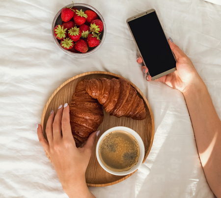 Woman using smartphone and having breakfast in bed. Feminine hands top view.の写真素材