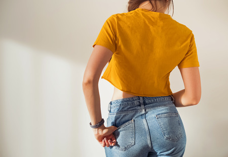 Young woman wearing yellow t-shirt and jeans posing against white wall, view from the backの写真素材