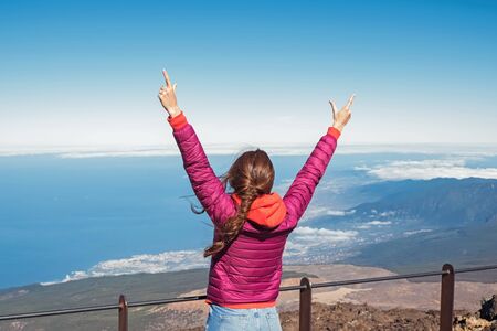 Young woman in pink jacket standing with her hands raised to the sky on the viewpointの写真素材